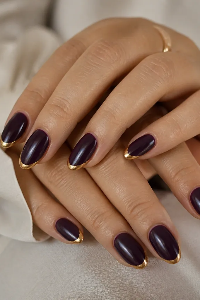 Short, almond nails with a glossy deep grape purple base. The tips are painted in a thin French line using antique gold, with a slight uneven foil texture at the edges.