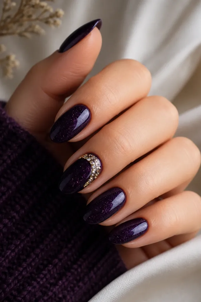 Dark purple nails with a glossy, slightly speckled night-sky finish. One nail has a gold gem crescent hugging the cuticle.