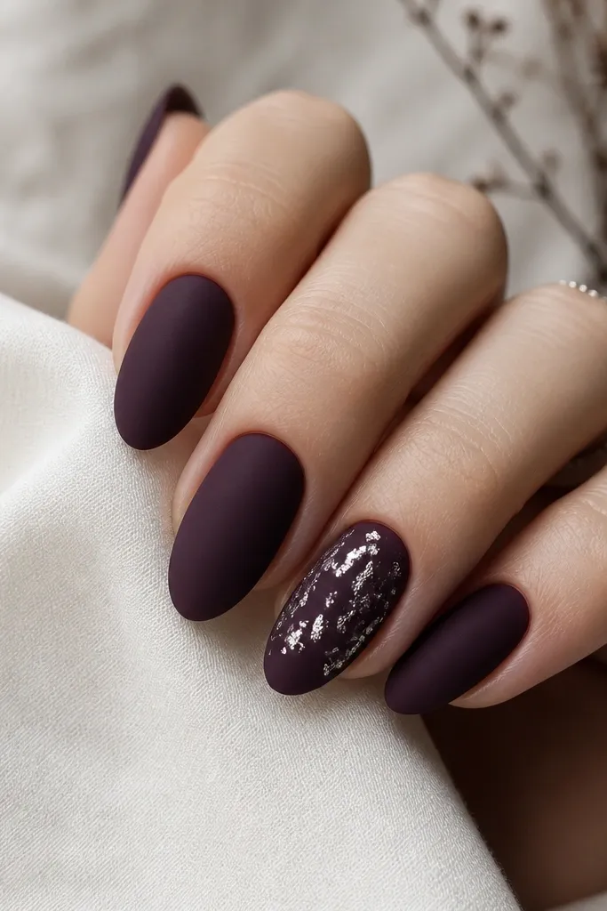 Matte dark purple nails with one accent. The accent has silver leaf pieces scattered like tiny flakes across the center and tip.