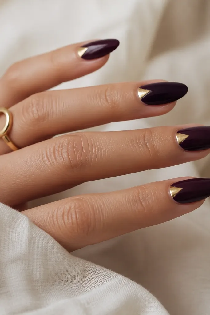 Deep purple nails with a sharp triangle cut-out near the sidewall. The triangle is filled with antique gold and creates a clean negative-space look.