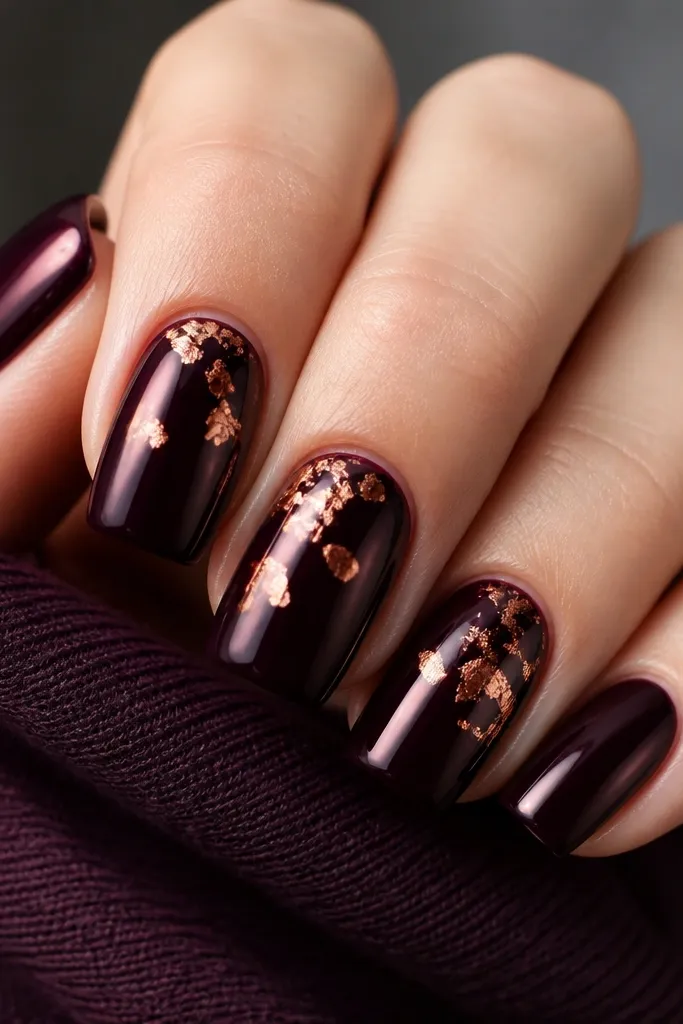 Square nails painted midnight plum, glossy and deep. On two or three nails, copper foil flakes sit near the cuticle and mid-nail like scattered metal dust. The flakes look irregular, with sharp copper edges catching light.