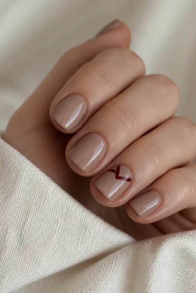 Short square nails in a mocha nude shade. On the ring finger, there's a small burgundy chevron pointing toward the center, with a glossy finish on all nails.