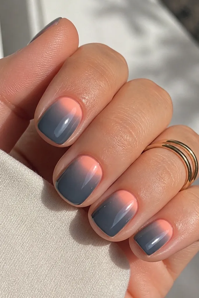 Short square nails painted charcoal grey with a peach-pink glow near the cuticle on each nail. The peach area fades into the grey and the nails are glossy.