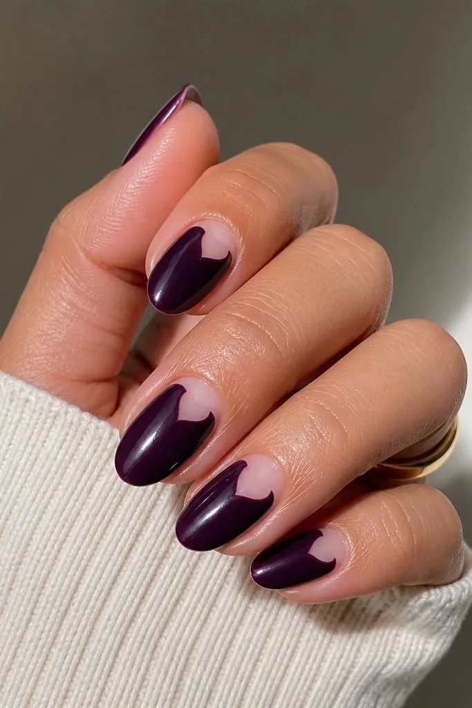 Short almond nails. A glossy dark purple covers most of the nail, but one side near the cuticle has a negative-space half-heart shape left clear. The half-heart is outlined by the purple edge.