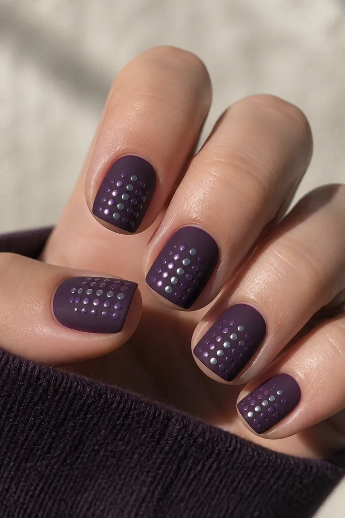 Short square nails. The base is matte dark purple. On each nail, there's a neat grid of tiny glossy dots in a lighter purple and silver, arranged in rows near the center and tip. The dots shine against the matte.