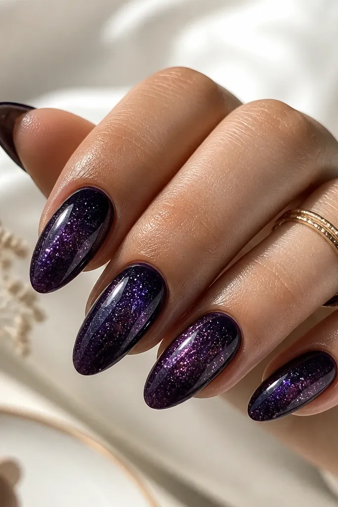 Almond nails with a deep dark purple galaxy effect. There are tiny specks like stars, concentrated near the center and fading toward the tips. Micro glitter sits softly over the gradient, with a glossy top coat.