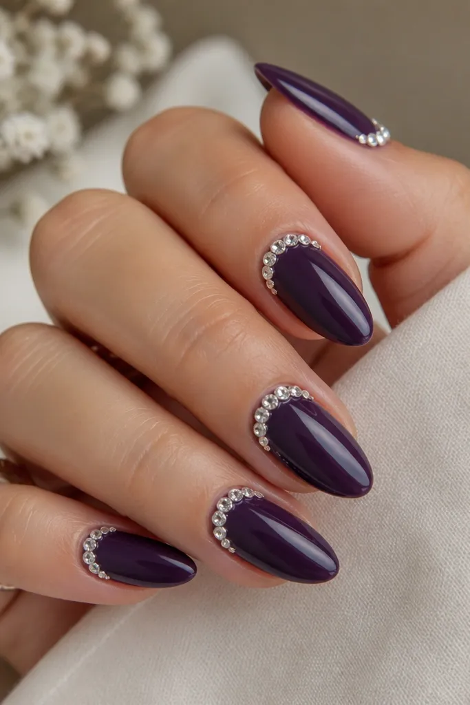 Medium almond nails with a solid dark purple base. On each nail, a crescent of small clear gems sits along one side near the cuticle, like a sideways tiara. The gems sparkle under a high-gloss top coat.