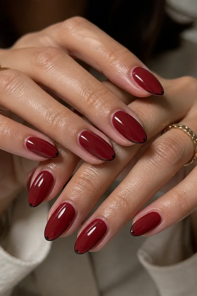 Short almond nails with a deep red base. A very thin black line runs along the smile line like a micro-French, and the very tip has a tiny black dot on each nail for balance.