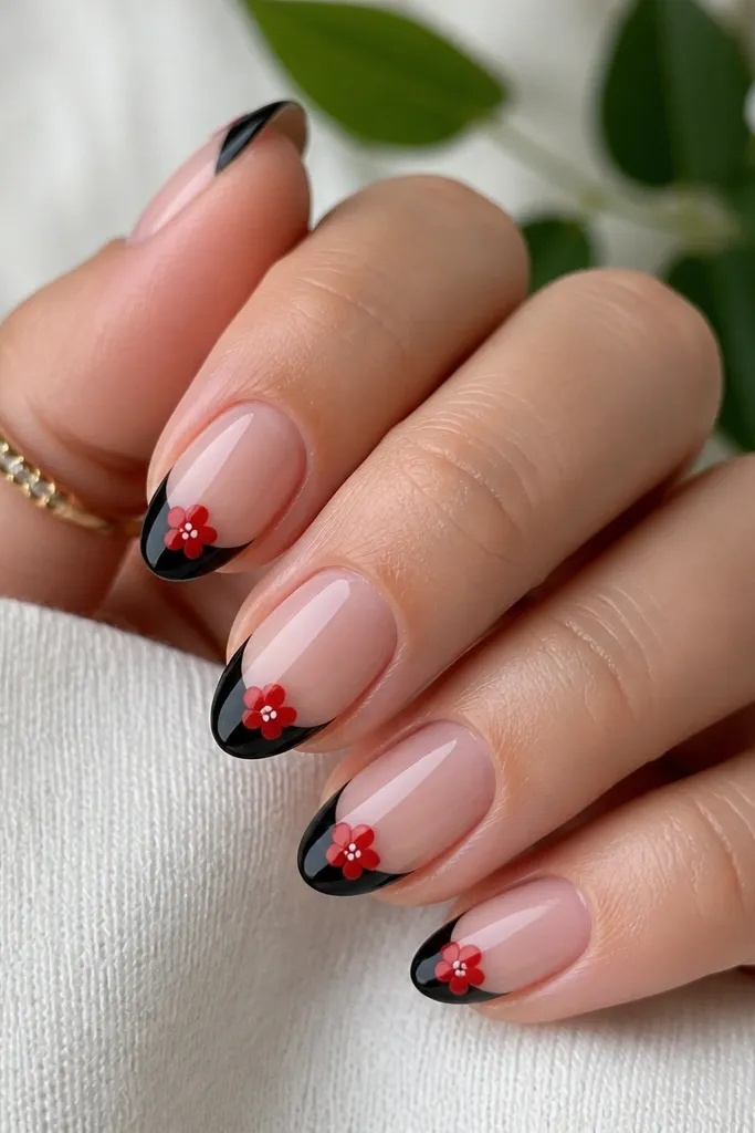 Short almond nails with deep black French tips. Each tip has one small red flower drawn near the outer side, with tiny white dot centers. The flowers look like simple 5-petal icons and the nails are glossy.