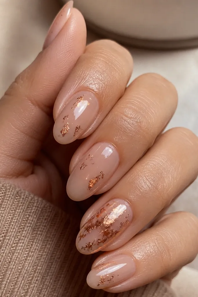 Warm spiced nude nails with scattered copper foil flecks, mostly concentrated on the ring fingers. The flecks are irregular and metallic, under a glossy topcoat.