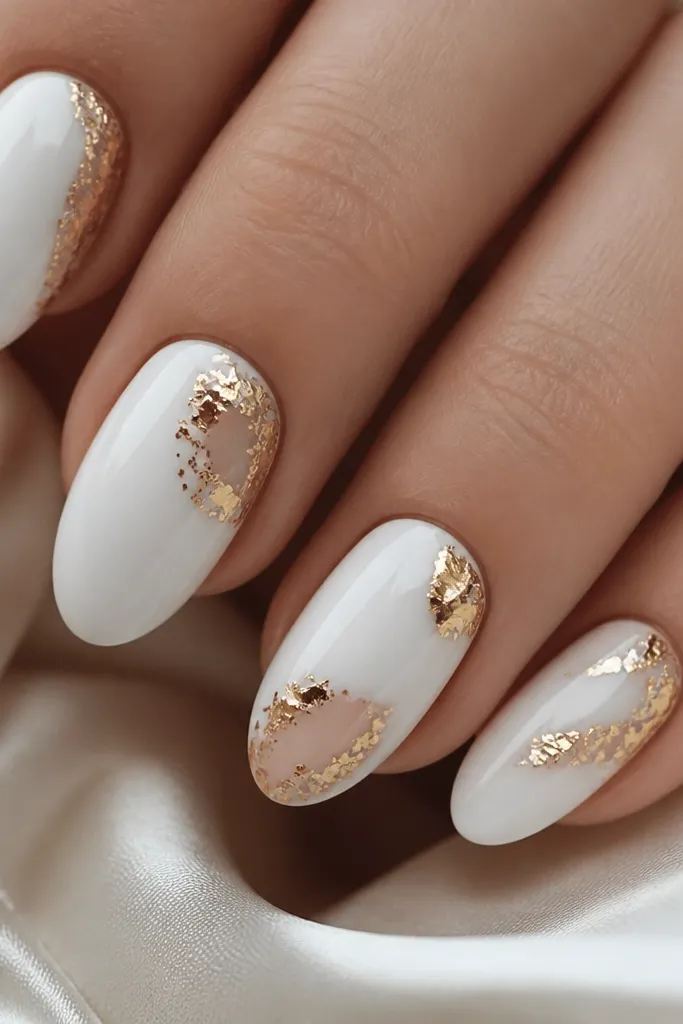 Nails with cool white on most of the nail but with small negative-space panels. Gold chrome leaf-like flakes sit on the panels and around the edges, with sharp metallic highlights.