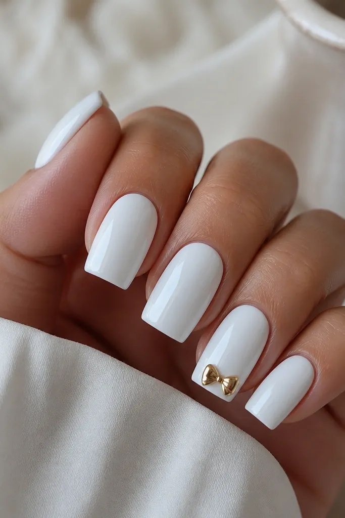 All nails are bright cool white. One accent nail has a small gold chrome bow centered near the tip, with two loops and a tiny knot, all mirror reflective.