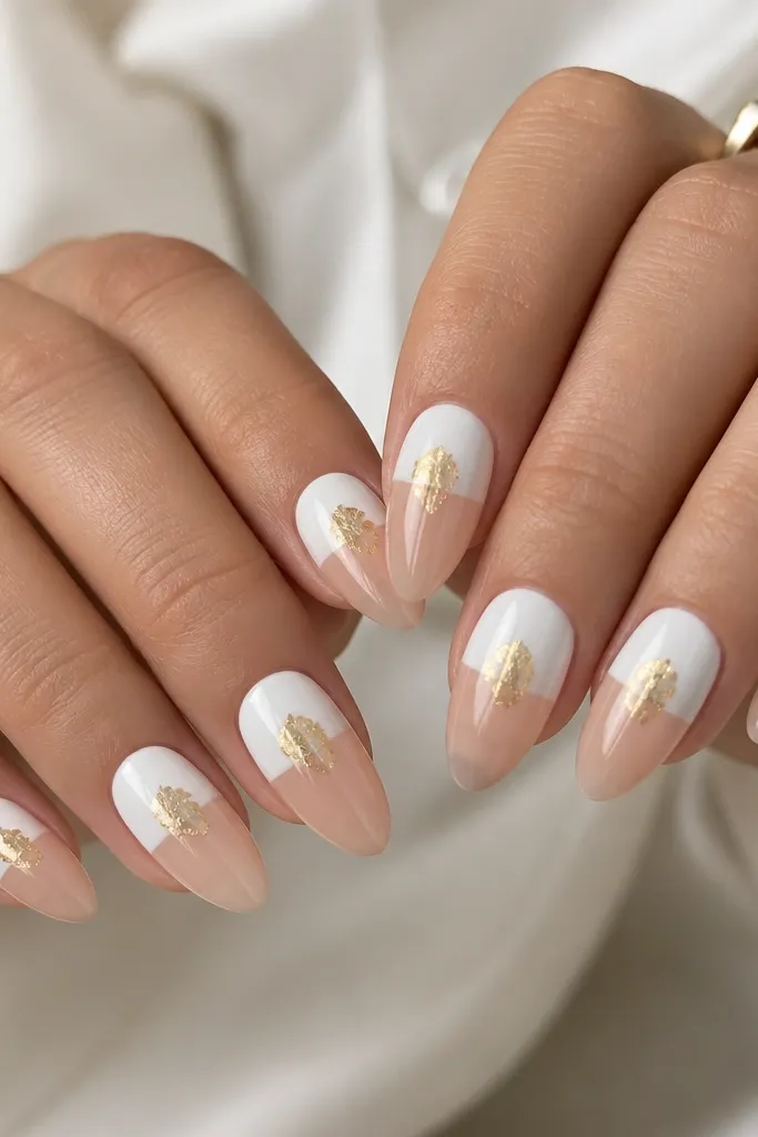 Almond nails with a nude base. The top half of each nail tip area is painted solid white, leaving the lower half nude. A small irregular gold foil splash sits in the white area near the center. Glossy finish, foil has irregular sparkle.
