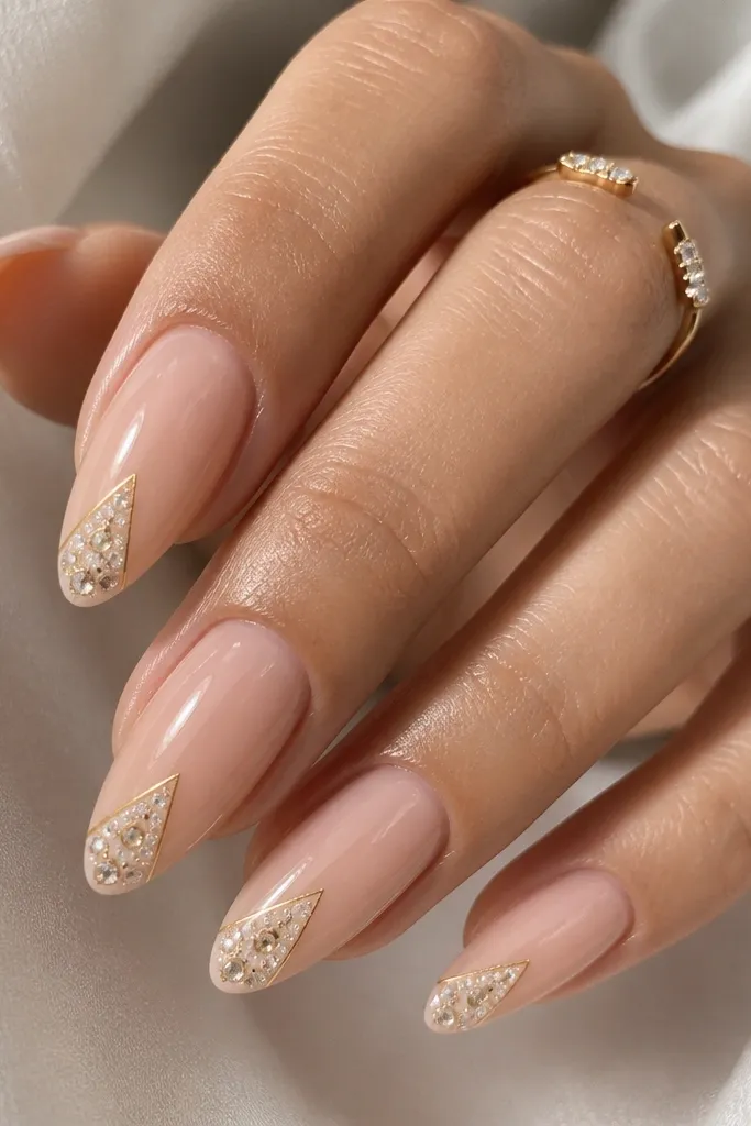 Almond nails with a nude base. Near the tip, there's a small white triangle filled area with tiny embedded studs or rhinestones. Around the triangle is a thin gold outline line. Glossy finish, studs sparkle.