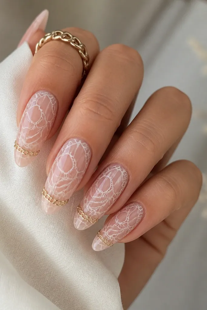 Almond nails with a sheer nude base and white lace-like lines. The lace pattern has negative space so nude shows through between white lines. Near the tip, there's a small gold chain link detail connecting two points. Glossy finish.