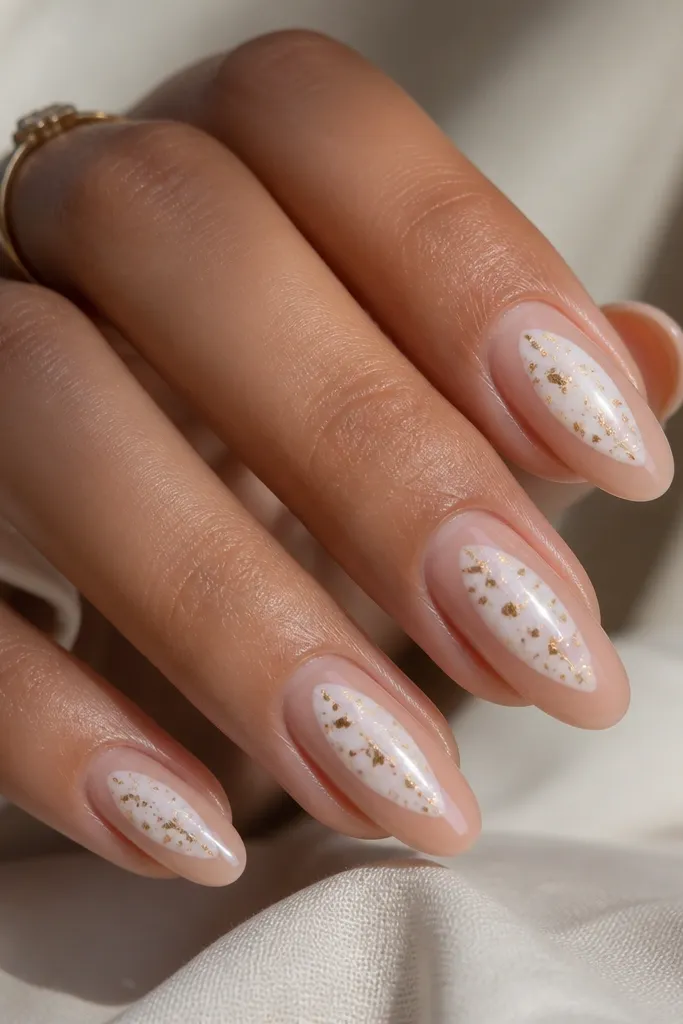 Nails have a sheer nude base with a white negative-space shape in the center. On top of the white area there are scattered tiny gold foil confetti pieces. Nails are glossy and the foil is sealed flat.