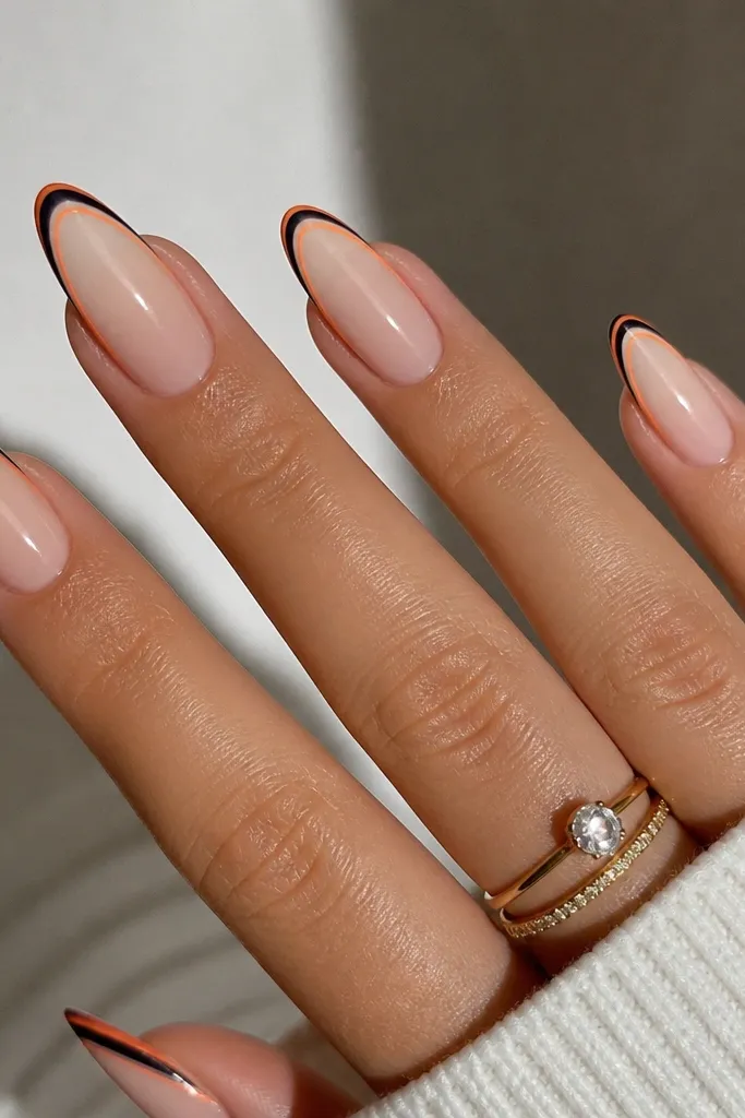 Long almond nails. The tips have a thin navy micro-French line. Around that navy line is a very delicate burnt orange outline, like a double border. The base is a sheer nude, and the tips are glossy.