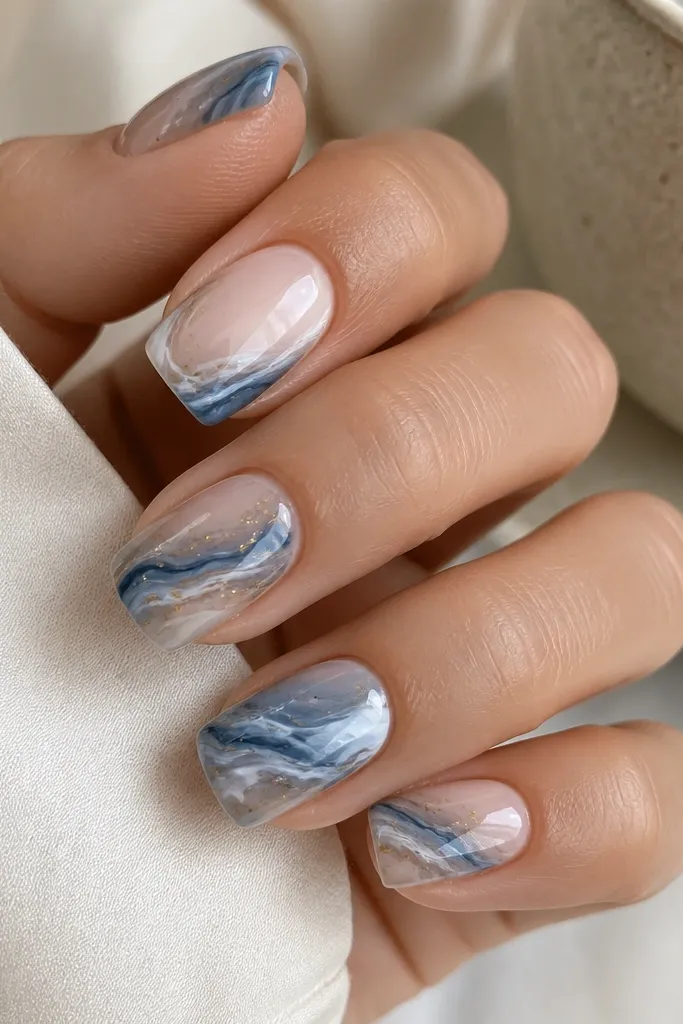 Square-to-squoval nails with swirling marble in blue-grey tones. The marble has organic white streaks and tiny gold flecks scattered like dust. The base is a soft cool nude, and the overall look is glossy.