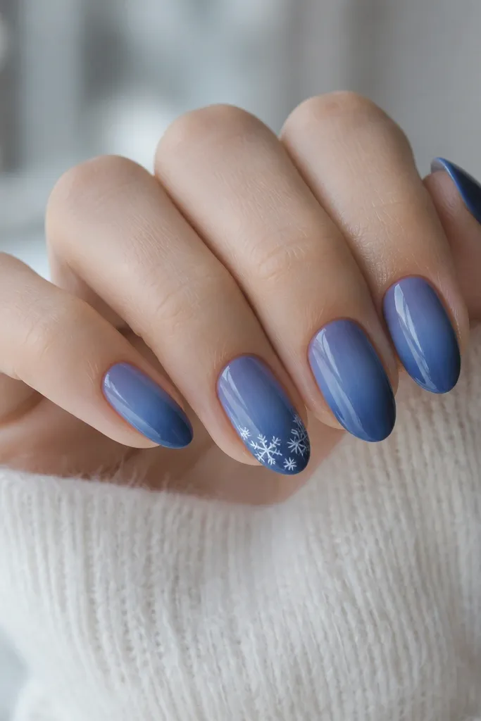 Short almond nails in indigo blue with a soft gradient from deep blue at the tip to lighter blue near the base. One accent nail has tiny white snowflakes clustered near the cuticle. All nails are glossy, with no other decorations.