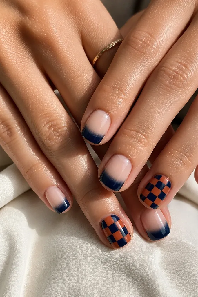 Short square nails. Most nails are sheer nude with a glossy navy shine at the tip like a soft gradient. Two accent nails have a checkerboard pattern made of navy squares and rust orange squares, with crisp edges. The accent nails stand out without extra glitter.