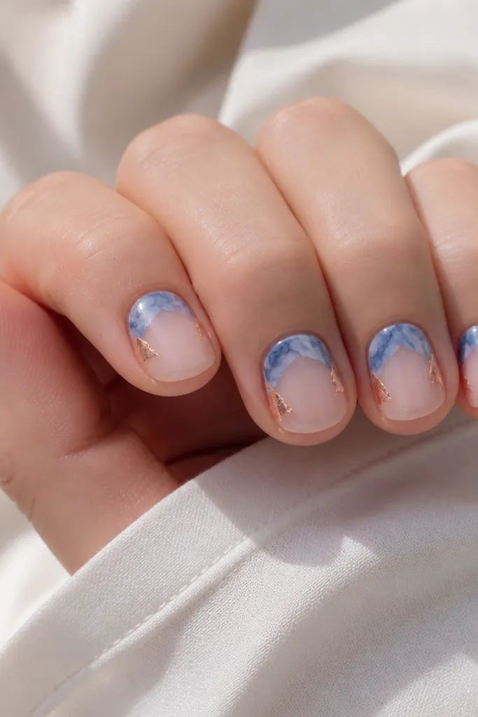 Short round nails. The base is sheer nude. The tips have blue marble patterns that occupy only the upper third. Two corners near the sidewalls on each nail have small copper foil pieces placed like tiny triangles.