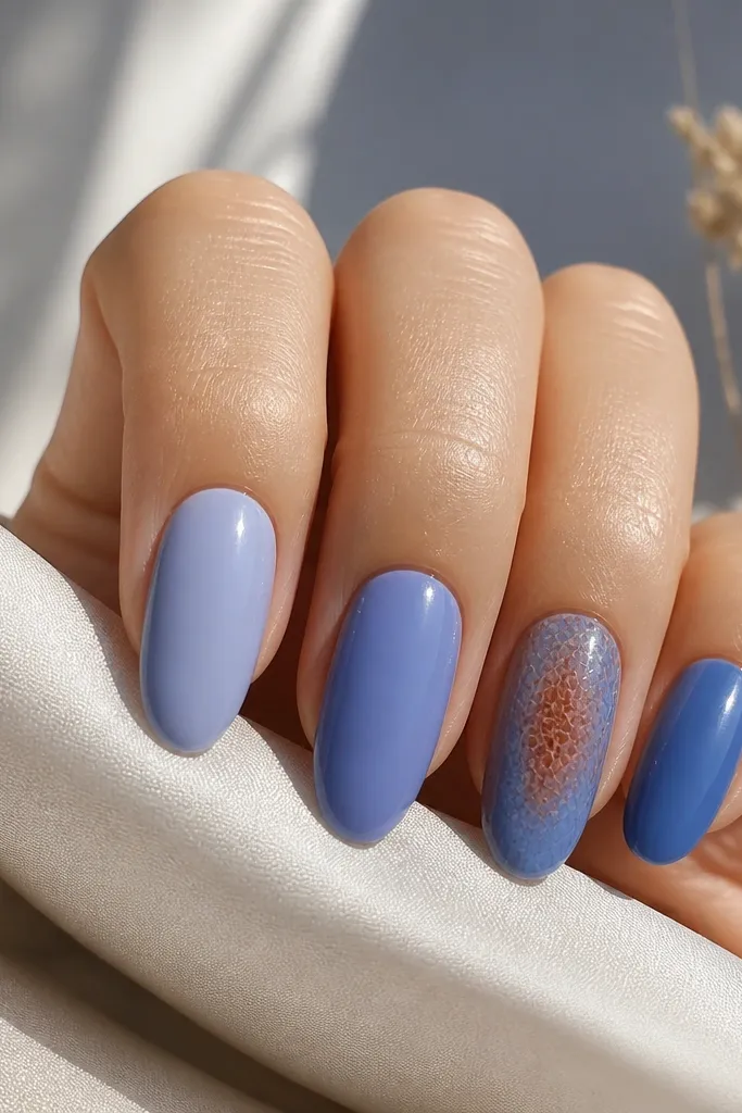 Five nails with a skittle gradient. Each nail shifts from periwinkle to a slightly deeper blue. The ring finger nail has a rust-brown "skin" texture over the center, like tiny scales, with a glossy top coat. Other nails are smooth and glossy.