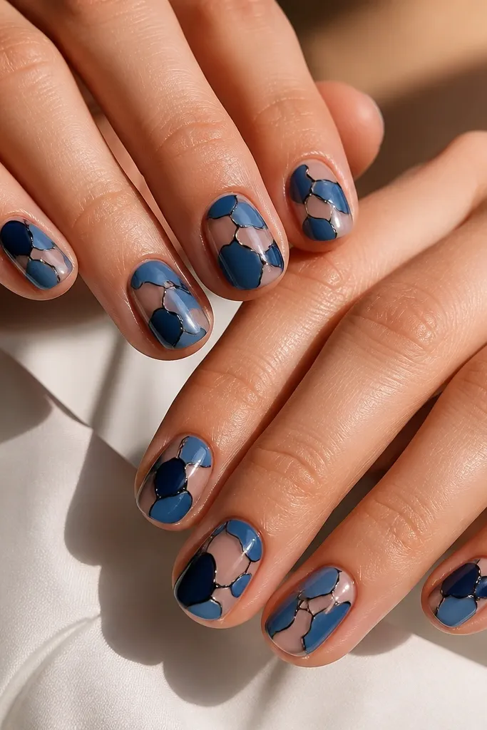 Short-to-medium nails with a warm nude base. Blue tortoiseshell patches sit across the nail in irregular shapes. Thin black lines outline each patch, creating a cell pattern. The finish is glossy, and the blue ranges from navy to medium blue.