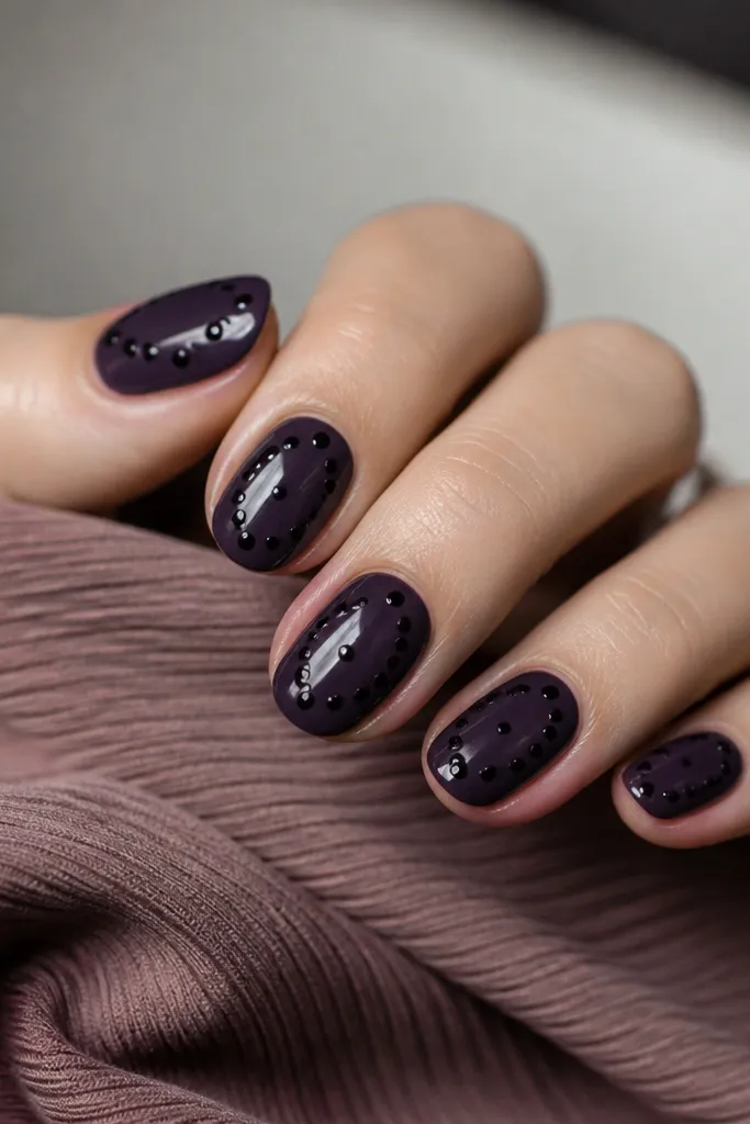Deep dark purple nails with tiny black polka dots scattered diagonally; glossy finish makes the dots look like raised ink.