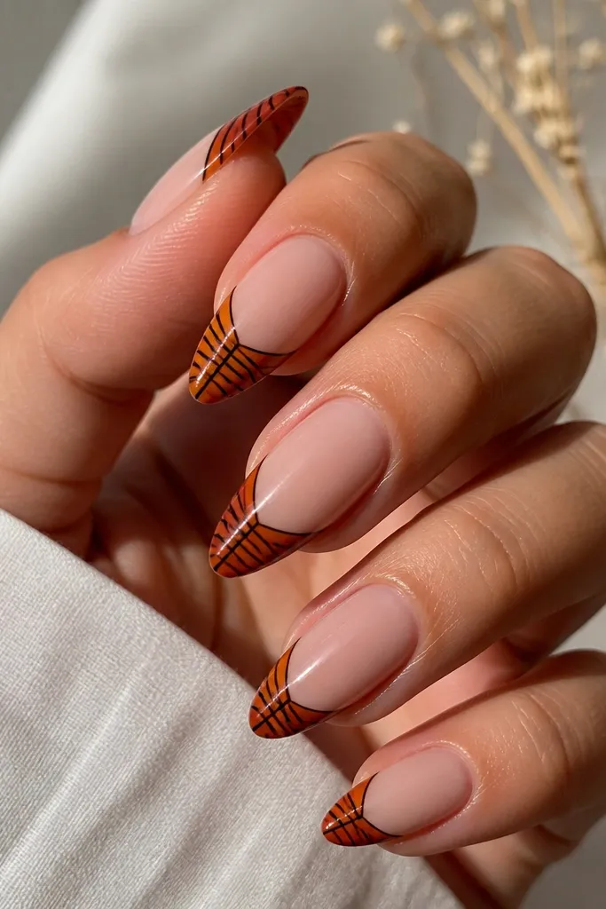 Almond nails with a nude base. The tips have a burnt orange band with thin black stripes running vertically and diagonally across the band. Glossy finish, sharp stripe lines.