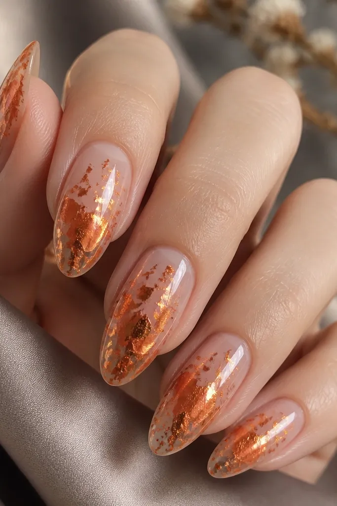 Clear nude nails with glossy finish. Rust orange foil flakes are scattered on the center and tip areas, with some flakes layered and others floating. The overall look is glossy and dimensional.