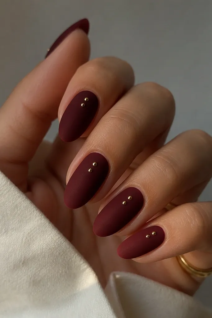 Deep oxblood nails in matte finish with one or two tiny gold studs near the center of each nail; clean almond shape, no other art.