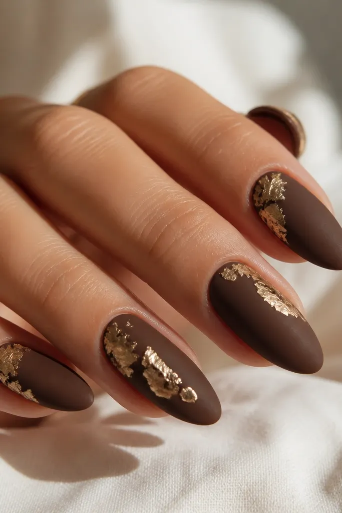 Matte chocolate brown almond nails with torn soft gold foil pieces placed at an angle near the cuticle and center; matte base with glossy foil highlights.
