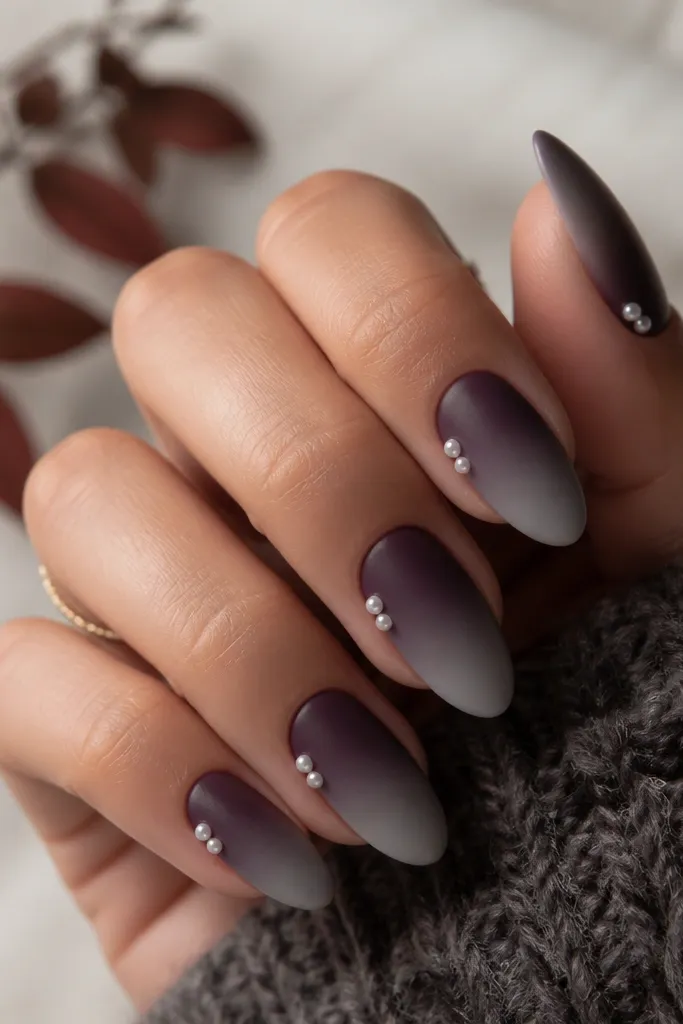 Autumn almond nails with a deep smoked plum base fading into a smoky smoke-gray ombré near the tips; two tiny pearl accents sit at the outer corners of each nail, catching light against a matte topcoat with a slight satin sheen.
