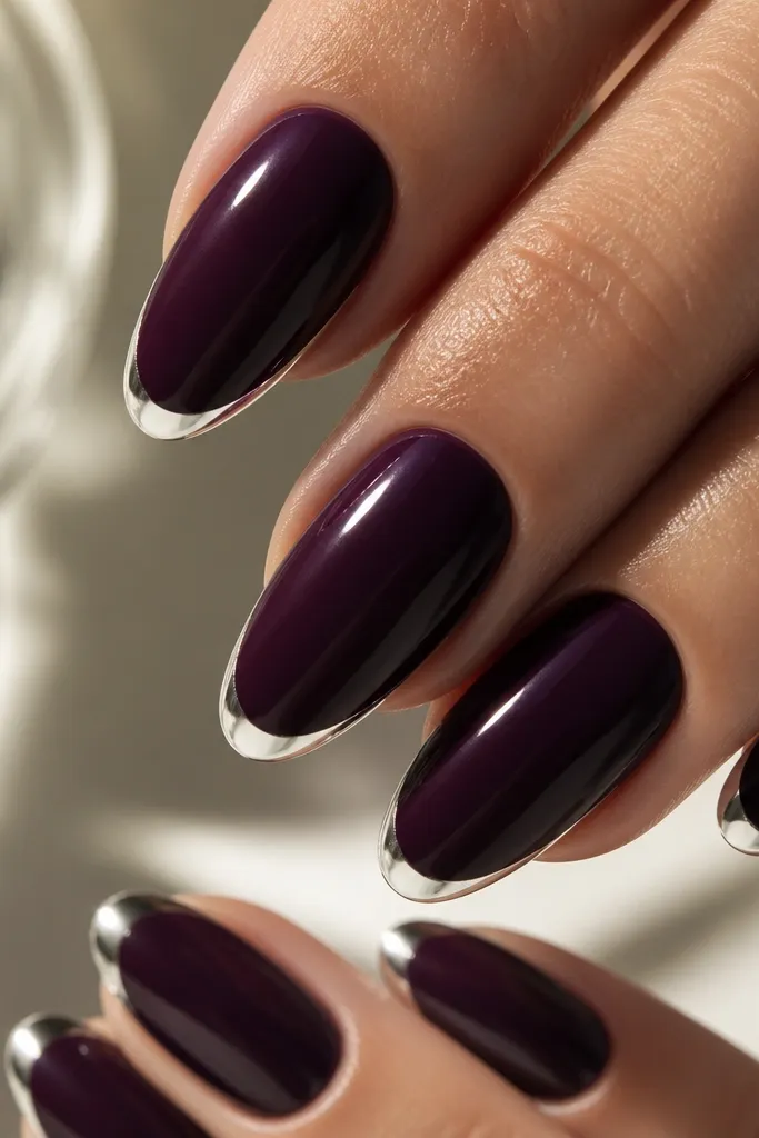 Close-up of almond nails painted a deep velvet plum. The tips have a thin, crisp silver arc only 1-2 mm wide, with a mirror-like shine. Cuticles are clean and glossy, no silver bleeding onto the purple.
