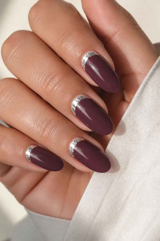 Medium almond nails in solid plum creme, glossy and smooth. A silver foil halo wraps the cuticle in a thin crescent, slightly irregular like real foil, with clean negative space around it.