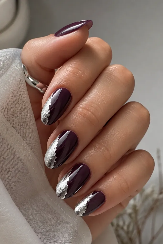 Medium almond nails painted deep plum. Silver foil wraps from the sidewall halfway across the nail, forming a diagonal metallic band. The rest of the nail stays solid plum and glossy.