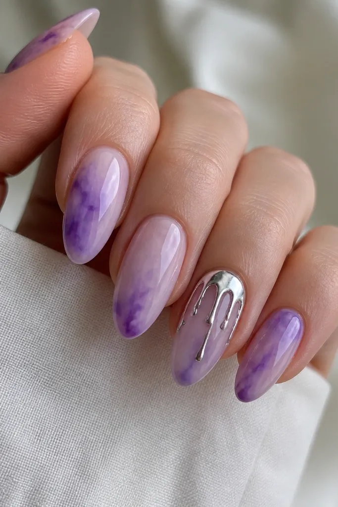 Almond nails with watercolor-style purple blends that look airy and smoky. One nail has a silver drip accent starting near the cuticle and tapering down, while other nails stay mostly solid. Glossy finish.