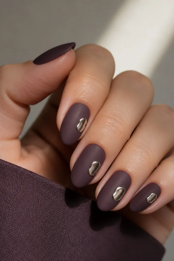 Short almond nails with a matte dark purple finish. On each nail, there's a small glossy silver patch near the center-right, like a metal sticker. The matte around it looks velvety.