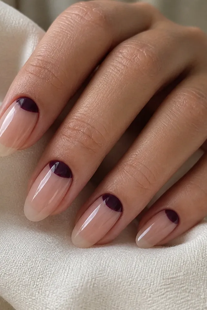 Medium almond nails with a sheer nude base; a deep grape reverse French half-moon at the cuticle; the rest of the nail is bare-gloss.