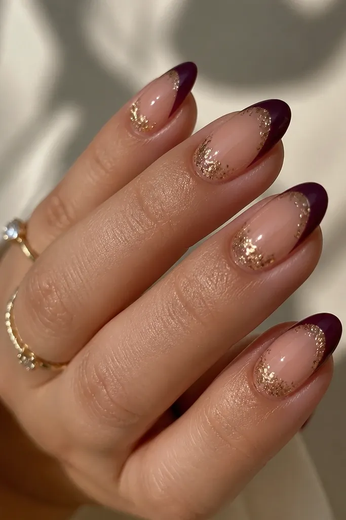 Almond nails with a sheer nude base; dark purple French tips; scattered tiny gold foil flecks concentrated near the smile line; glossy top.