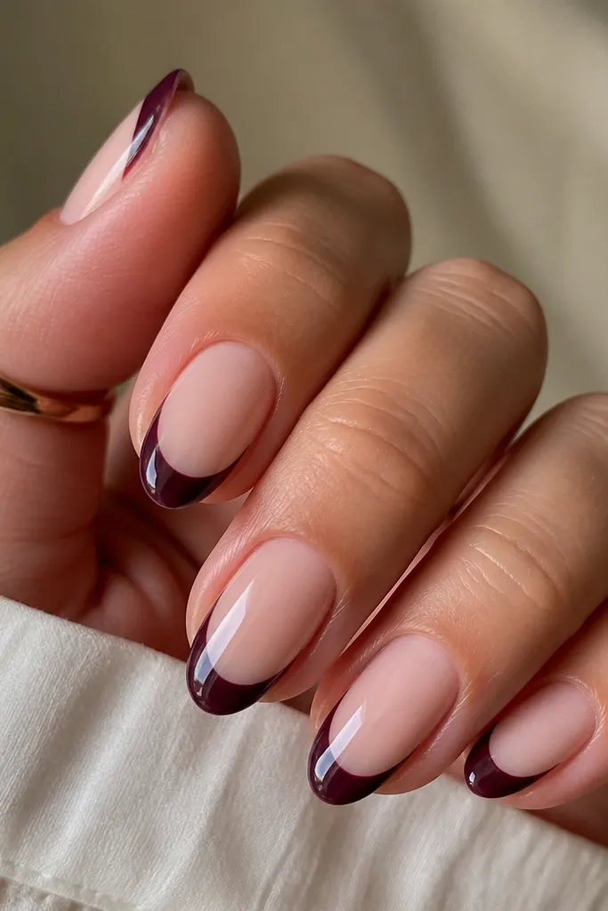 Short almond nails with sheer nude base; dark plum French tips that leave thin negative-space cutouts near both sidewalls; glossy finish.