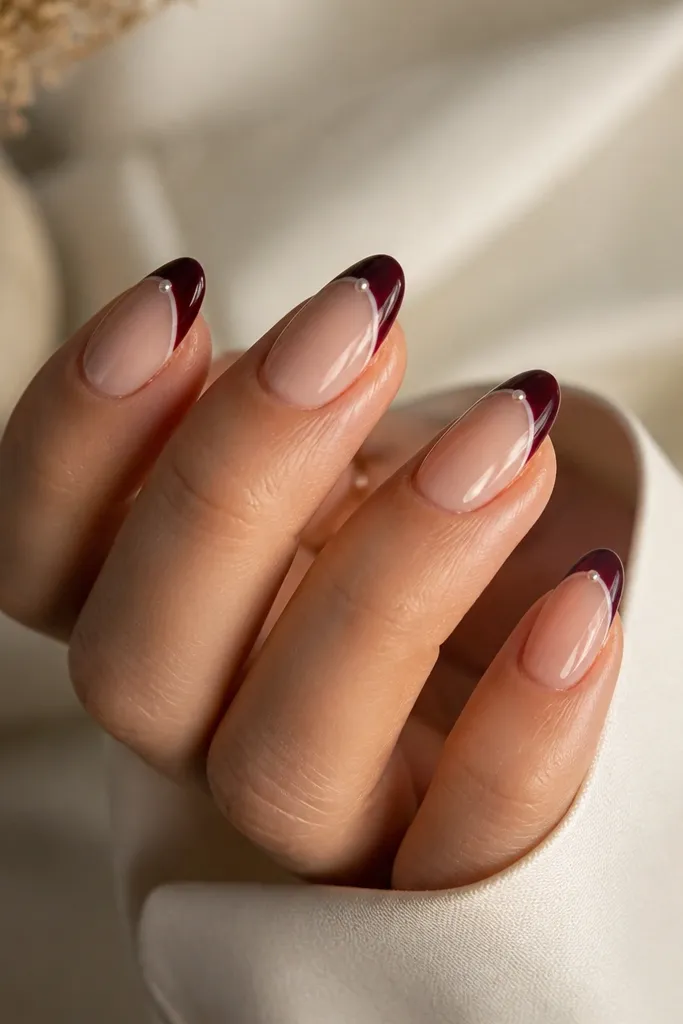 Almond nails with nude base; dark grape French tips; one small pearl dot centered on each tip near the smile line; glossy finish.