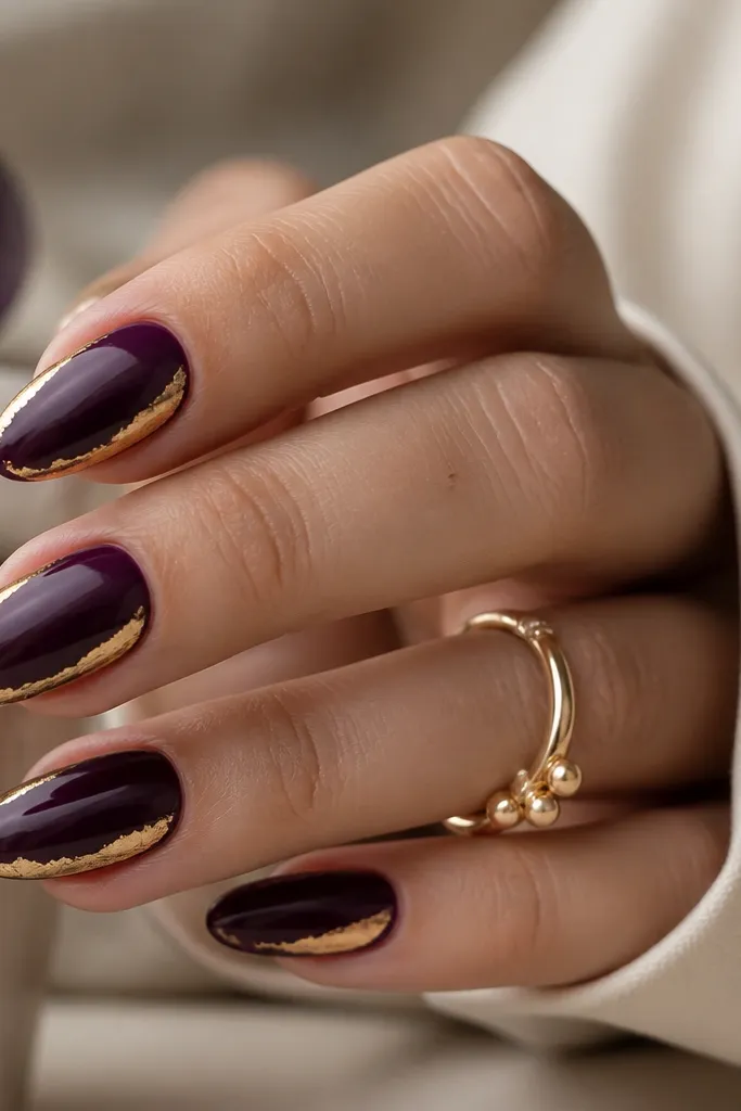 Glossy dark purple almond nails with thin strips of gold foil placed along the side edges near the tip, creating a delicate frame effect.