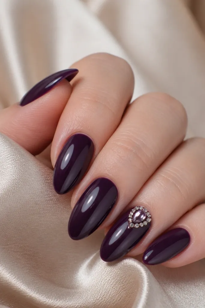 Dark purple glossy almond nails with one nail featuring a single teardrop rhinestone near the cuticle, surrounded by tiny clear crystals on the same nail.