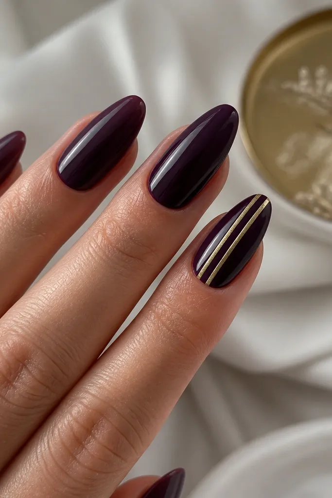 Glossy dark purple almond nails with one accent nail showing a thick gold stripe and a thinner parallel stripe, separated by a small gap, placed diagonally from side to center.