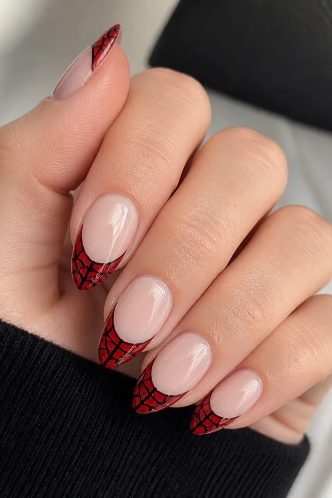 Medium almond nails. Most nails are sheer pink nude. The tips are painted glossy red like a French manicure. Over the red tips, black spider web lines radiate from near the center, with fine crackle gaps between lines.