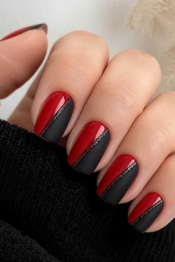 Short squoval nails. Each nail has a diagonal split: left side glossy red, right side matte black. Along the diagonal seam is a thin line of fine black micro-glitter that catches light. One accent nail has both colors but the glitter line is thicker at the center.