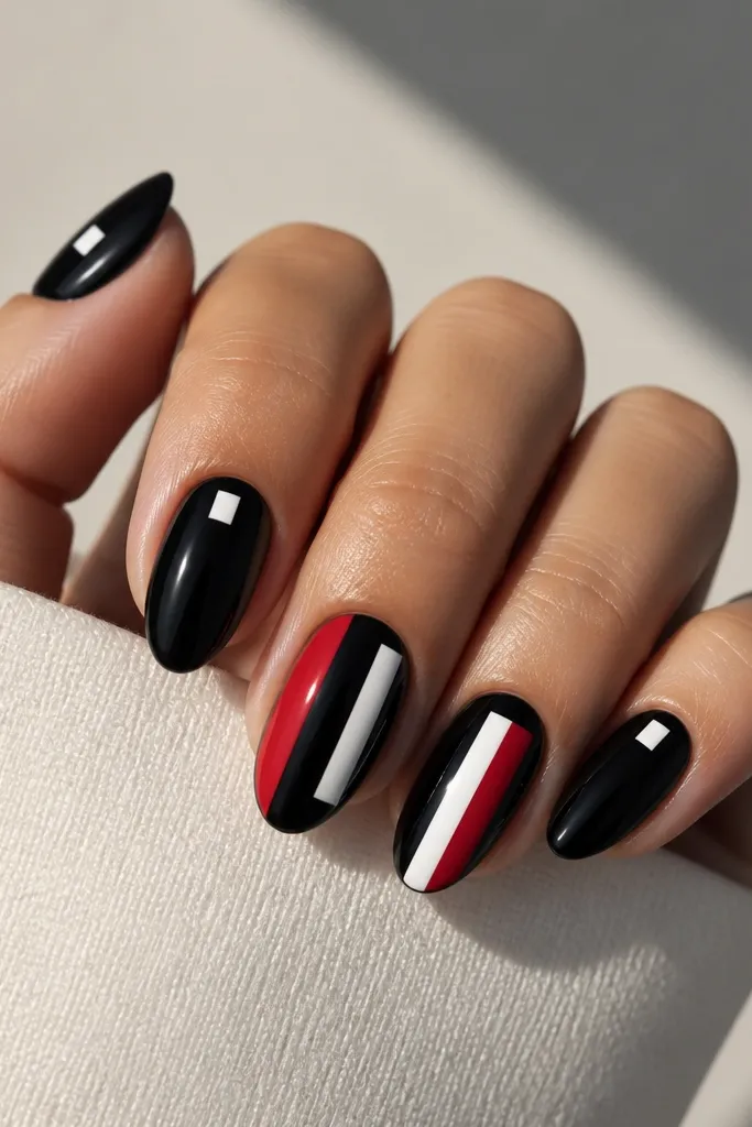 Medium almond nails with a glossy black base. Two nails have vertical blocks: a red rectangle on one side and a white rectangle on the other, separated by black space. Other nails are solid black with one small white block.