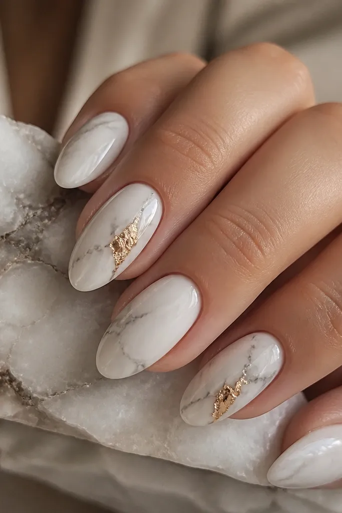 Milky white nails with soft gray-white marble veining. On two accent nails, small foil gold pieces sit where the veins meet, like gold trapped in stone. Everything is glossy and smooth.