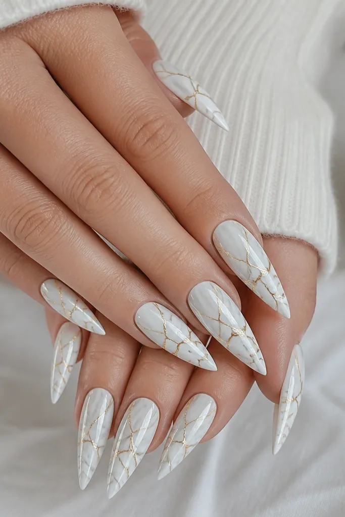 Stiletto nails with white marble swirls in soft gray. Thin gold veining lines cross the marble like cracks, concentrated around the middle and tapering toward the tip.